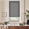 Patterned gray roller blind hangs lowered over a kitchen window, covering most glass above a sink; countertop holds bowls of leafy greens, potted plant, and framed art on the wall.