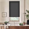 Dark gray roller blind covers most of a window above a kitchen sink; stainless faucet, wooden cabinets, countertop with bowl of greens, potted plants, and framed sketch on the wall.