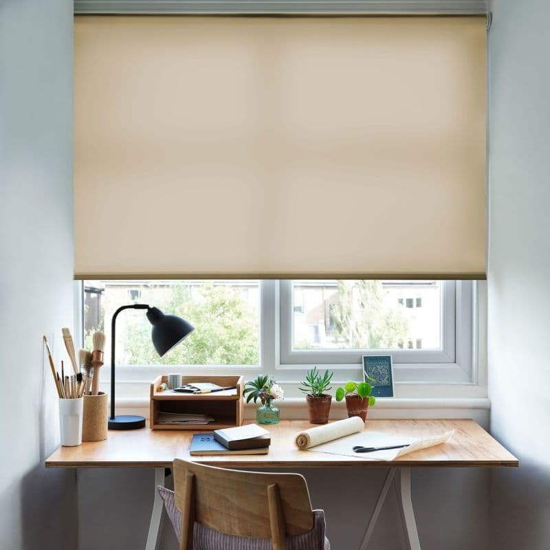 Beige roller blind lowered nearly fully, diffusing daylight through its fabric; covers a double window above a tidy wooden desk with lamp, notebooks, brushes and small potted succulents.