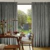 Floor-to-ceiling pleated grey patterned curtains, hung in soft folds and parted centrally; drawn partly open, admitting daylight and a garden view; wooden dining table set for breakfast beneath a pendant lamp.