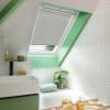 White pleated skylight blind, lowered halfway across the roof window; partly closed, filtering soft daylight; compact attic bathroom with green trim, white herringbone tiles, sink and bathtub.