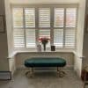 White plantation shutters cover a bay window, louvers partially tilted, filtering daylight; bright sitting alcove with a green velvet bench on gold legs and a vase of red flowers.