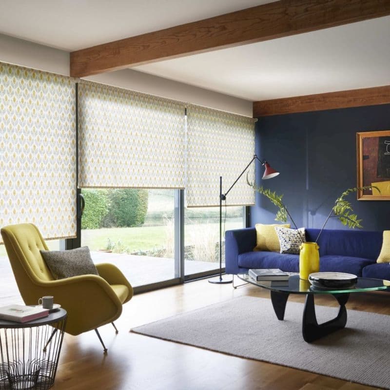 Patterned roller blinds covering floor-to-ceiling sliding doors, pale yellow geometric leaf print, lowered and filtering daylight; modern living room with navy sofa, yellow armchair, exposed wooden beams and garden view.
