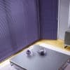 Purple horizontal blinds covering corner floor-to-ceiling windows, tightly closed with visible pull cords; blocking light. Modern living room with low grey coffee table, purple glass ornaments, hardwood floor, grey sofa.