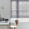 Sheer grey Roman blind, lowered across a multi-pane window with horizontal folds, filtering soft daylight; minimalist home office with white desk, wooden chair, pendant lamp, and bare tree branches outside.