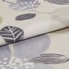 A folded beige curtain fabric with stylized gray and lavender leaf prints (textured knit), overlapping to show the hem and pile, photographed close-up on a neutral surface under soft lighting.