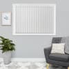 White vertical blinds fill a rectangular window — evenly spaced slats. They are fully closed, softly diffusing daylight. Neutral grey living room with charcoal armchair, potted plant, and framed print.