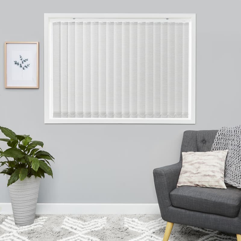 White vertical blinds fill a rectangular window — evenly spaced slats. They are fully closed, softly diffusing daylight. Neutral grey living room with charcoal armchair, potted plant, and framed print.