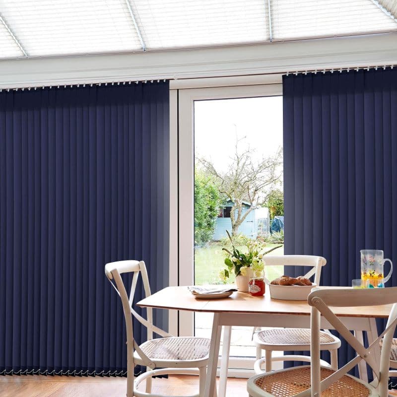 Vertical navy blinds, ceiling-mounted, form closed side panels with a central gap, blocking light; a dining table and chairs occupy the room, garden visible through the glass door.