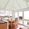 Pale honeycomb/pleated blinds covering the sloped conservatory roof and windows, closed and softly diffusing daylight; small dining area with oak table and beige chairs beside a kitchen counter, patio view.