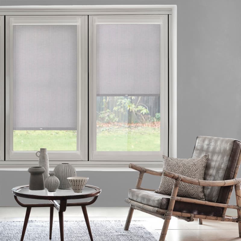 Striped pleated cellular blinds covering twin windows, one fully lowered and the other partially lowered, filtering diffuse daylight; modern gray living room with wooden armchair, side table, and backyard view.
