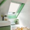 A white pleated skylight blind, partially lowered and filtering daylight over a roof window; set in green-trimmed sloped ceiling above a tiled bathroom with sink, tub, plant, and toiletries.