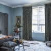 Patterned heavy curtains and sheer Roman blinds — curtains parted and pooling at the floor, blinds partially lowered filtering soft daylight — in a softly lit blue bedroom with rug, bench, and stacked books.