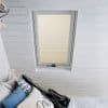 Beige pleated skylight blind, fully closed and filtering daylight, set inside a white-framed roof window; attic bedroom with white painted wooden walls and a bed piled with patterned cushions.