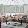 Light-green pleated cellular blinds lowered halfway across tall bay windows, filtering soft daylight; conservatory-style living room with beige sofa, wooden coffee table, blue rug, floor lamp and decorative cushions.