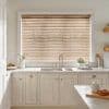 Wooden horizontal blinds (light oak slats, pull cords) mostly closed over a window, filtering daylight into a bright neutral kitchen with a double sink, marble counters and potted herbs.