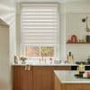 Beige dual-layer zebra roller blind, lowered covering most of the window, alternating opaque and sheer horizontal stripes filtering daylight; modern kitchen sink area with brass faucet, wooden cabinets, marble countertops.