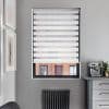 Zebra-style roller blind, lowered with alternating opaque and sheer horizontal bands, partially revealing bottom pane; filtering daylight in gray kitchen corner with coffee machine, radiator and framed prints.