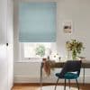 Light-blue fabric Roman blind, lowered to show a narrow bottom gap and horizontal folds; filtering daylight over a small desk with chair, mirror, vase of flowers, and neutral walls.