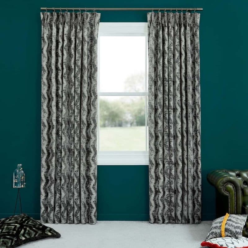 Black-and-white wavy-striped pleated curtain panels, hung on a metal rod and parted, framing the central window and admitting daylight into a teal-walled room with cushions and a green leather armchair.