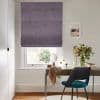 A textured purple Roman blind, fully lowered with horizontal folds, filters daylight over a sash window; a wooden desk, teal chair, vase of flowers and framed seaside print sit below.