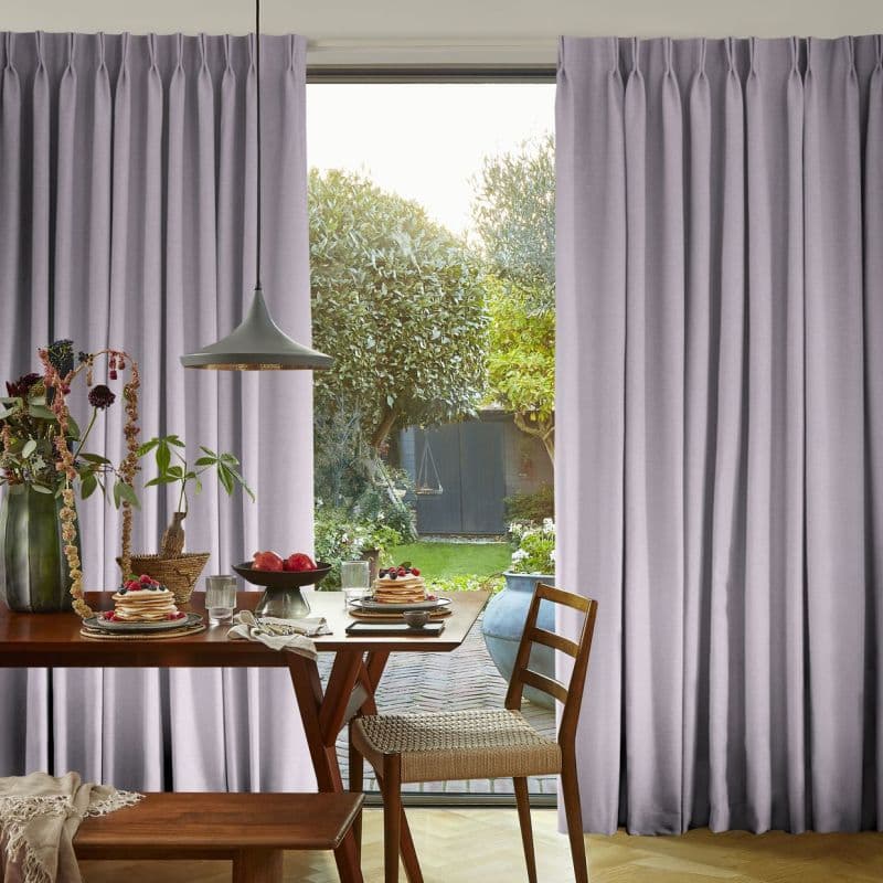 Floor-length pleated lavender curtains, drawn slightly apart to reveal a central opening, softly filtering daylight; dining table with pancakes and pendant lamp in foreground, backyard garden visible beyond.