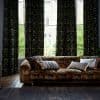Heavy dark green patterned curtains, mostly drawn with central gaps, filtering daylight through tall bay windows; in front, a low patterned sofa with cushions on wooden floor, brick view beyond.
