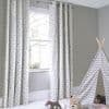 Grommet-top curtains with grey star print, hanging full-length on a metal rod, filtering bright daylight through the window into a softly lit grey nursery with a chevron teepee and toys.