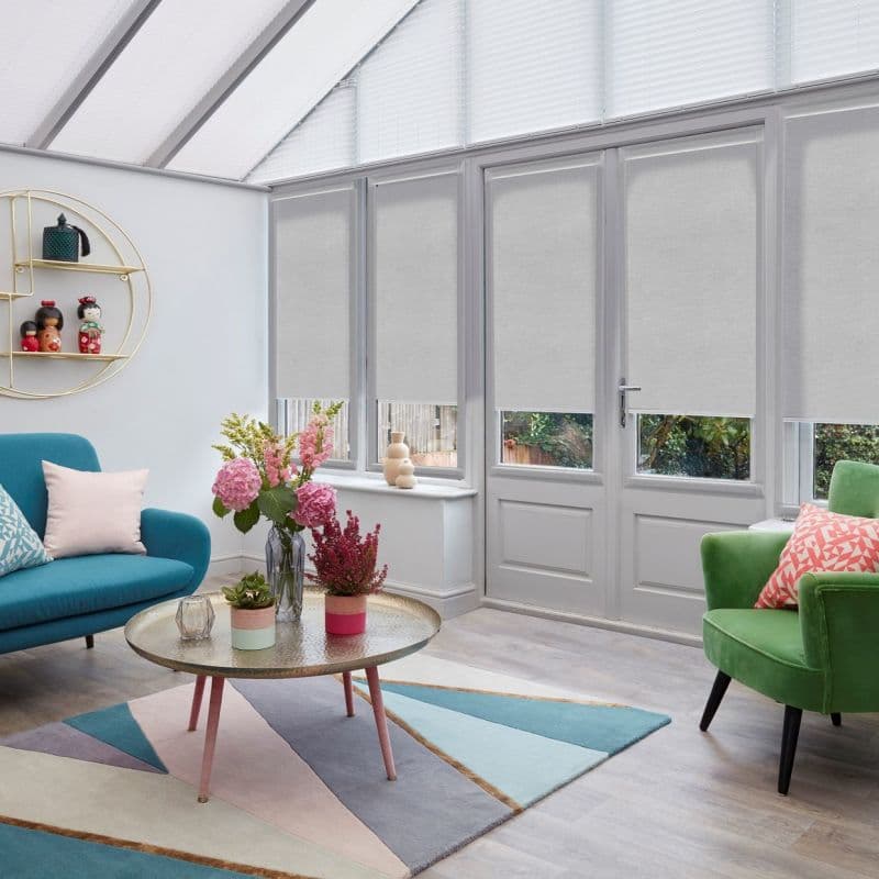 Light-grey roller blinds cover tall glazed doors and windows, partially lowered to leave narrow bottom panes clear, filtering daylight in a bright conservatory living area with pastel furnishings.