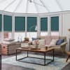 Teal pleated cellular blinds, fitted inside white conservatory window frames and lowered, filtering daylight; in a cozy conservatory living area with a beige sofa, pink knit throw, and wooden coffee table.