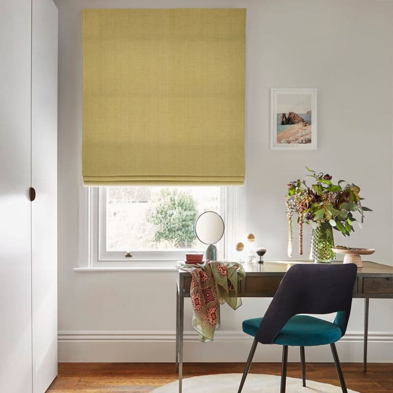 Pale mustard fabric Roman blind, fully lowered, filtering daylight through a small lower gap; above a wooden desk with teal chair, vase of flowers, framed print and garden view.