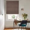 Brown fabric Roman blind, fully lowered with a narrow light gap, textured finish; filtering daylight over a wooden desk with chair, mirror and vase of flowers in a sunlit home office.