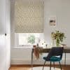 Beige-and-white diamond-patterned Roman blind lowered over a window, filtering soft daylight; a desk beneath holds a mirror, vase of flowers, scarf and teal chair in a bright, neutral home office.