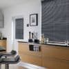 Grey horizontal Venetian blinds covering a large window and adjacent door window, slats tightly closed dimming light; in a modern kitchen with light-wood cabinets, black countertop, bar stools, small appliances.