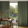 Floor-length, pleated olive-green curtains, drawn open at the center, framing a sliding glass door; they reveal a sunlit dining corner with a wooden table set for breakfast and a leafy backyard.
