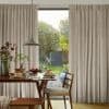 Beige floor-length pleated curtains, hung and parted at center revealing a glass door, filtering soft daylight onto a wooden dining table set for breakfast, with a green garden visible beyond.