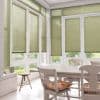 Green pleated window blinds, fully lowered across tall French doors and surrounding windows, lightly filtering sunlight; dining room with white wooden table and chairs, floral cushions, tile floor, garden view.
