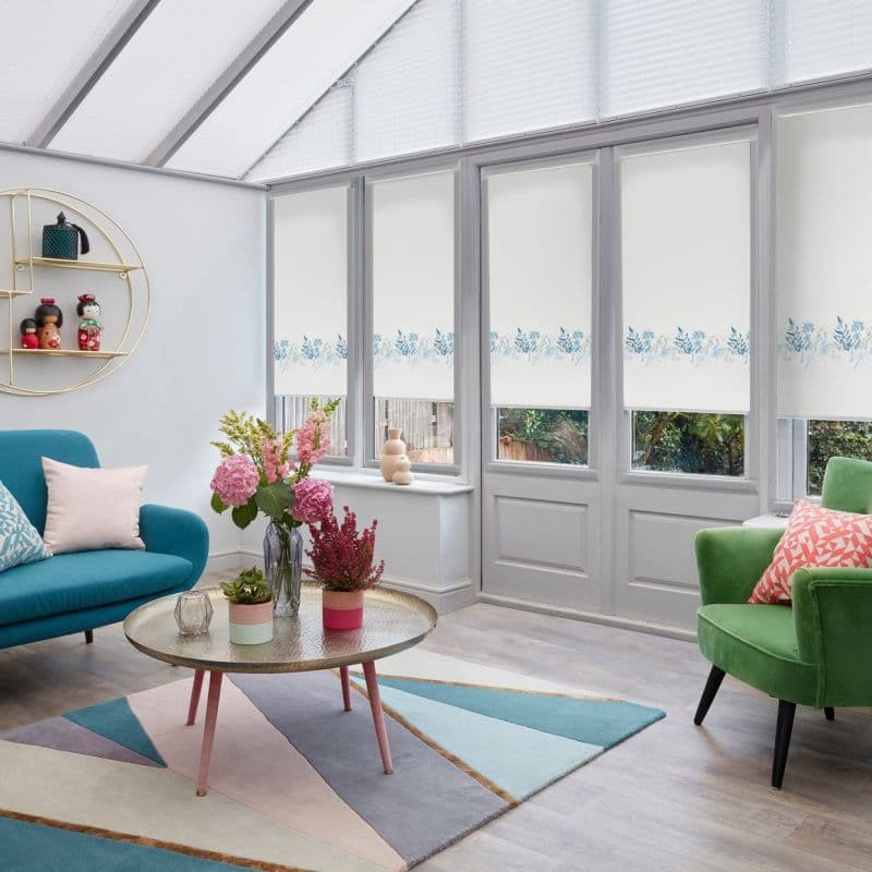 White roller blinds with a blue floral border, lowered across tall glass doors and roof windows, filtering daylight in a conservatory with sofa, armchair, coffee table and a garden view.