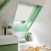 White pleated skylight blind, partially lowered across an angled roof window, filtering daylight — bathroom with green-painted window trim, white herringbone tiles, bathtub, sink, potted plant, and toiletries.