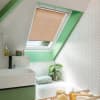 Beige pleated skylight blind, lowered to cover most of a roof window and filtering daylight; in a sloped, green‑accented bathroom above a bathtub with white herringbone tiles, plant, sink.