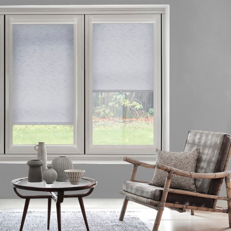 Light-gray roller blinds cover twin tall windows — left fully lowered, right half-raised — softly filtering daylight over a living room with a wooden armchair, small table with ceramics, and a grassy garden view.