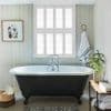 White plantation shutters covering a tall window, slats closed and filtering bright daylight; set above a black clawfoot bathtub in a light‑paneled, softly lit bathroom with towel and potted plant.