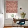 A terracotta, abstract-patterned Roman blind lowered over a sash window, softly filtering daylight; above a desk with a teal chair, vase of flowers, round mirror, and pale walls.