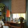 Brown Roman blind with white diamond pattern, lowered and filtering daylight over a window; set above a wooden desk with lamps, books, sculptures, teal walls and houseplants in a study.