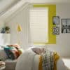 White horizontal blinds covering a window, closed and filtering daylight; in an attic bedroom with sloped ceiling, yellow wall accent, bed with colorful pillows and a yellow floor lamp.