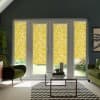 French doors fitted with yellow floral roller blinds partially lowered, filtering light into a modern living room with green armchairs and sofa, glass coffee table on chevron rug, potted plants.
