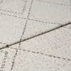 Beige woven rug with thin dashed dark grid lines, folded at the edge to show layered thickness and looped texture; close-up studio shot with neutral lighting on a flat surface.