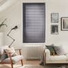 Gray horizontal Venetian blinds covering a tall central window, closed and filtering soft daylight; slender radiator beneath; neutrally furnished living room with armchair, sofa, framed art, and lamp.