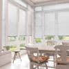 White horizontal Venetian blinds lowered across tall bay windows, slats mostly closed and softly filtering daylight, in a bright dining sunroom with white table, chairs, cushions and garden view.