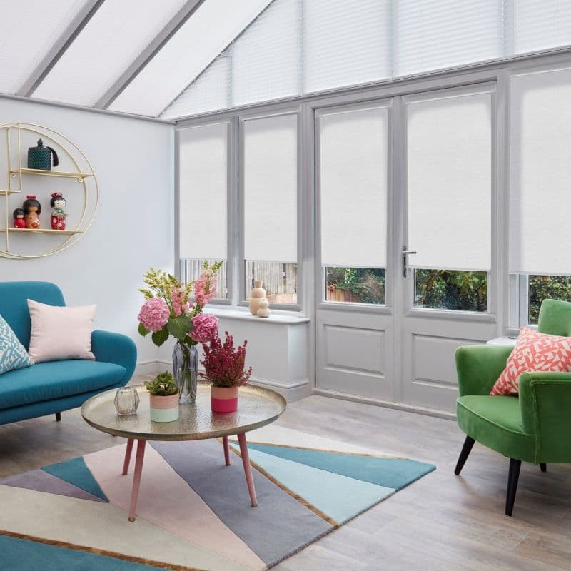White roller blinds lowered across tall glass doors and lower windows, filtering daylight; triangular pleated blinds above. Context: bright conservatory-style living room with colorful sofas, coffee table and plants.
